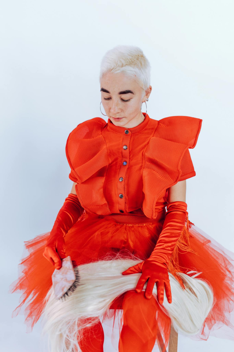 Fashion-forward woman in red outfit brushing a blonde wig, showcasing style and creativity.